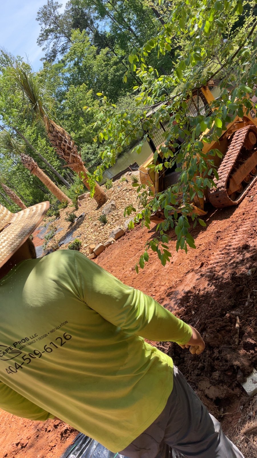 Landscaping site prep with tracked loader, graded red dirt, and palm trees on a gravel slope