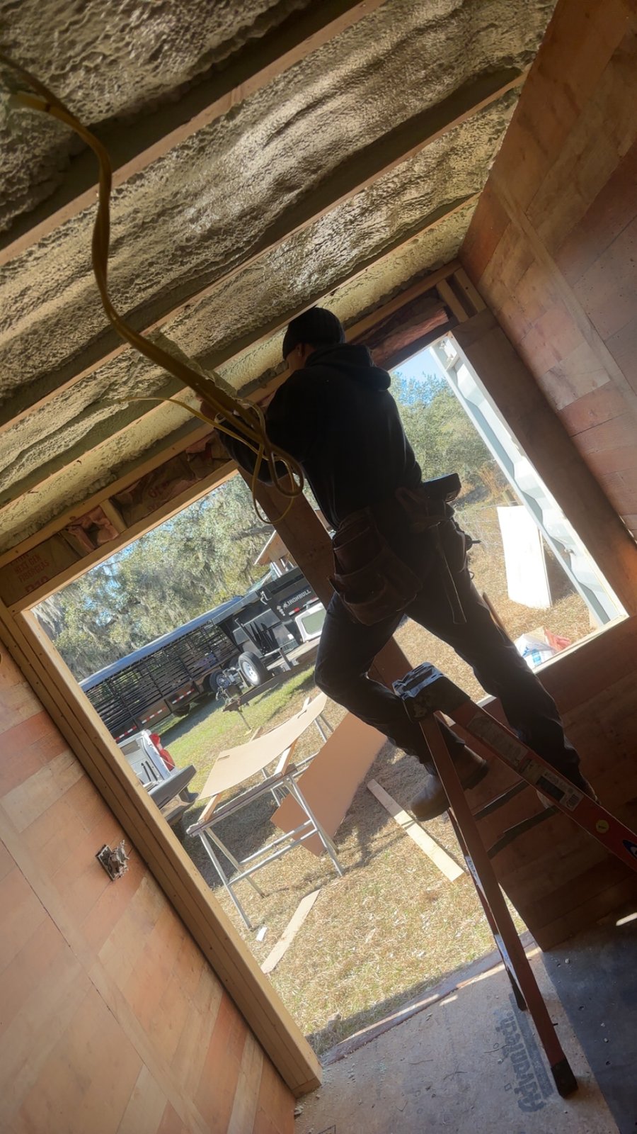 Worker on a ladder applying spray foam insulation in a wood-paneled room